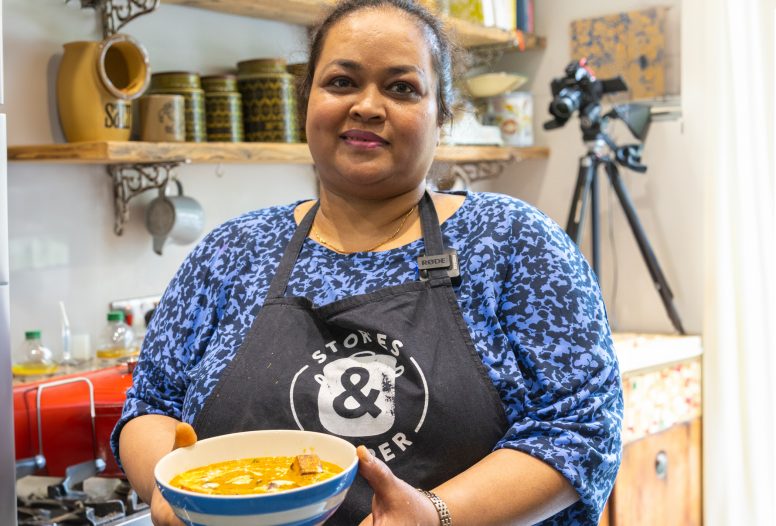 Shahnaz wears a stories and supper apron, smiles at the camera and holds a bowl of the paneer curry she has just made.