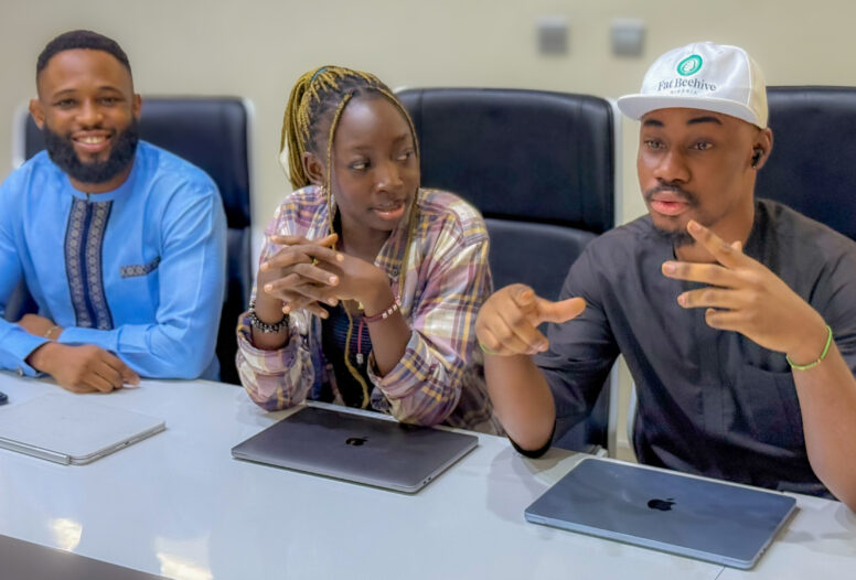 Three people sit at a conference table with laptops, having a discussion. One man in a blue shirt smiles, a woman in plaid listens attentively, and another man in a hat gestures while speaking.