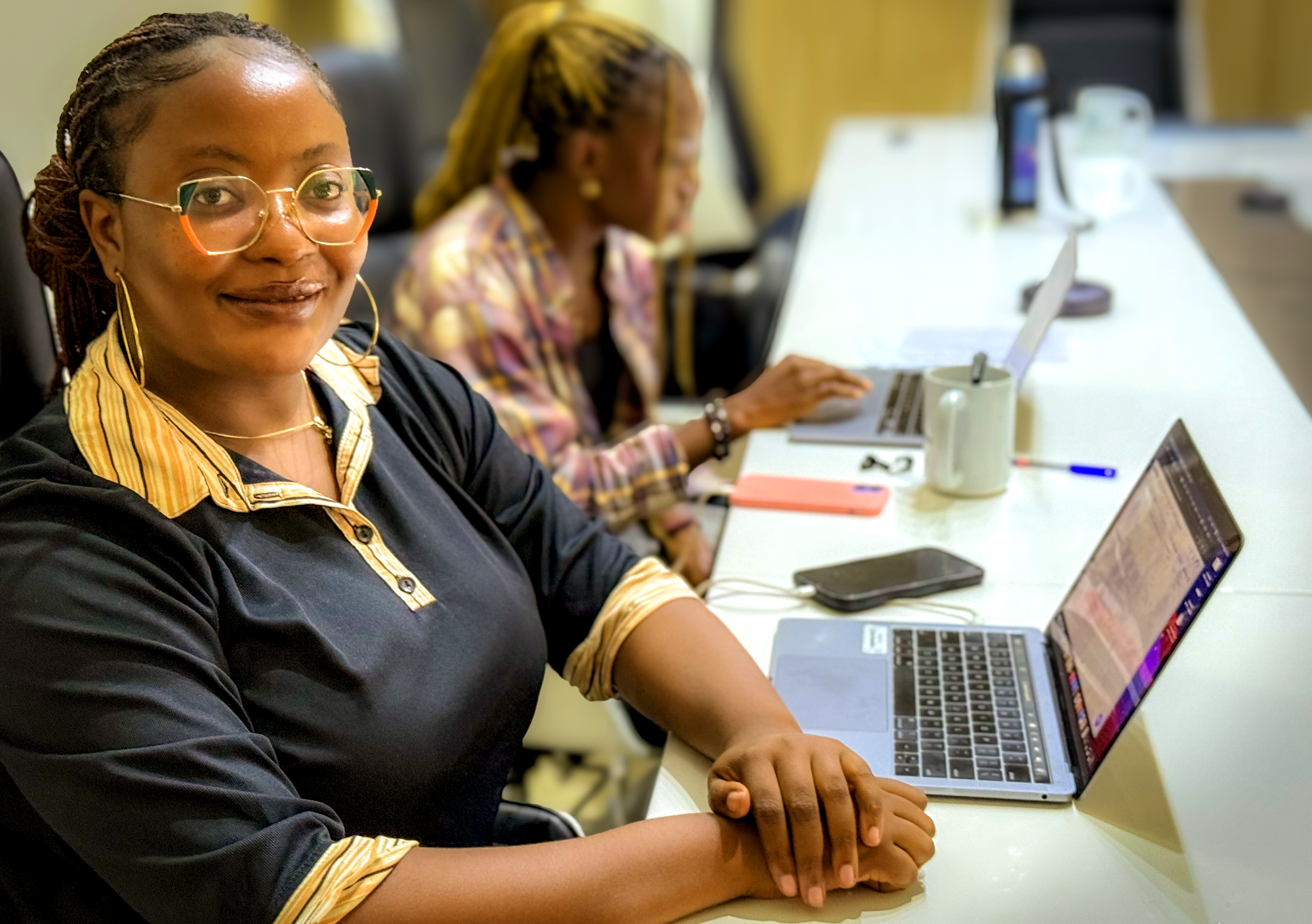 A Black woman with braided hair and distinctive orange and green glasses smiles at the camera from an office desk with a laptop and phone. Another woman works in the blurred background.