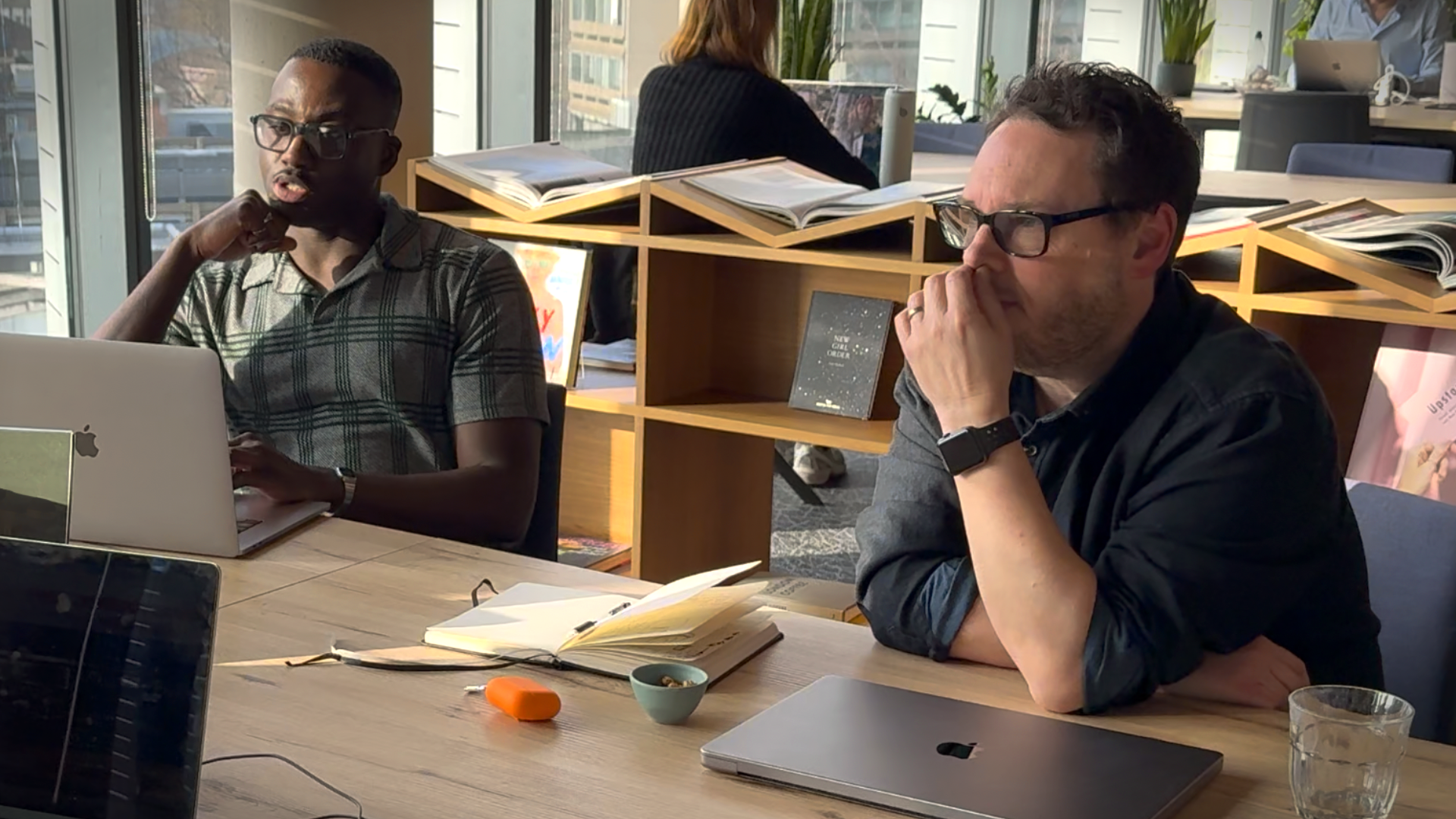 Two men working at a wooden table in a bright office. One man types on a laptop, while the other listens thoughtfully with his hand to his face. Laptops, notebooks, and snacks are on the table.