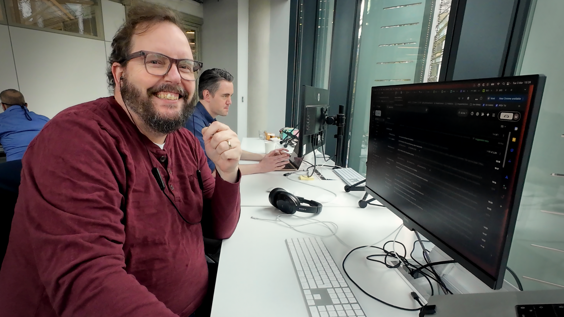 A man with a beard, glasses, and a maroon shirt smiles broadly at the camera from an office desk with computer monitors and keyboards.