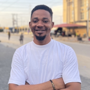 Joshua Adumchimma smiling with arms crossed, wearing a white t-shirt, standing outdoors on a street.