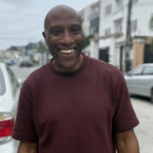 A bald man wearing a maroon t-shirt smiles broadly while standing on a residential street with parked cars and buildings in the background.