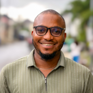 A young man with a short beard and black-rimmed glasses smiles confidently outdoors, wearing a green striped zip-up polo shirt with a blurred street scene in the background.