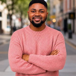 A smiling young man with a beard stands with his arms crossed on an urban street, wearing a pink knit sweater, with blurred city buildings and trees in the background.